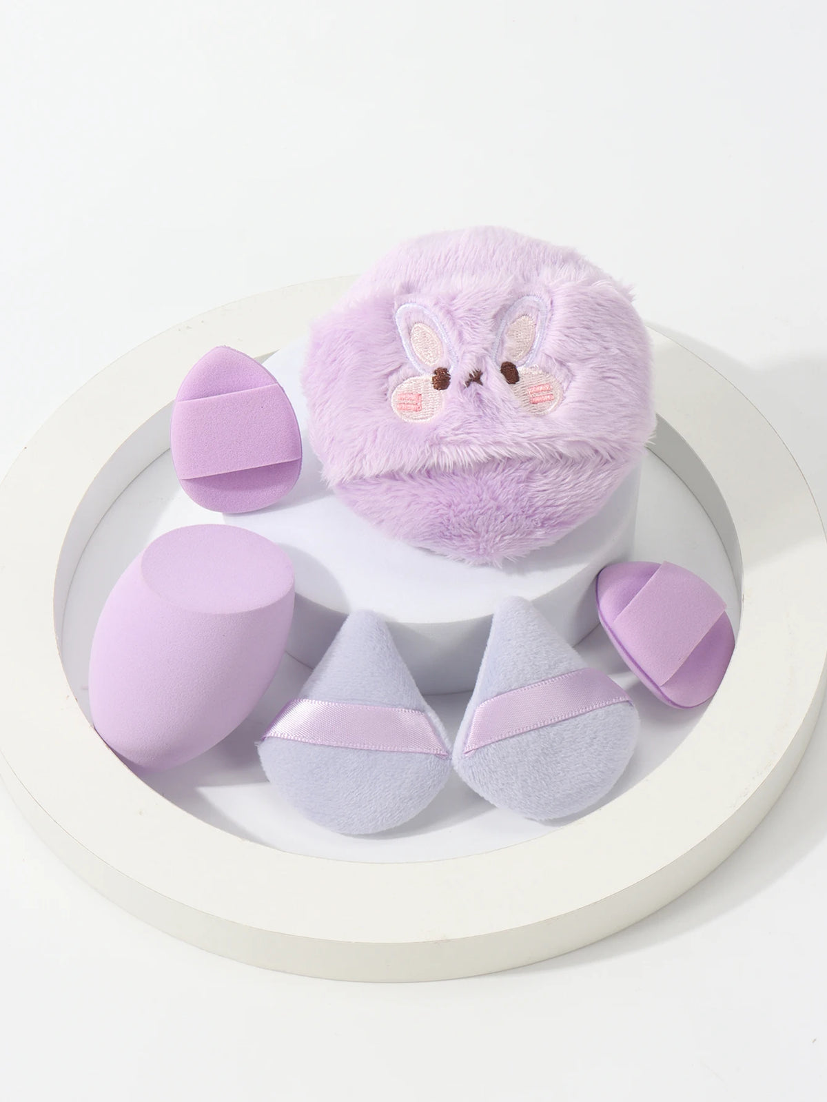 Un set de poudres légères lavande, comprenant un gros puff fluffy et plusieurs petits poudres roses et violets.