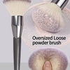 A silver oversized loose powder brush with soft purple and beige bristles shown next to a hand.