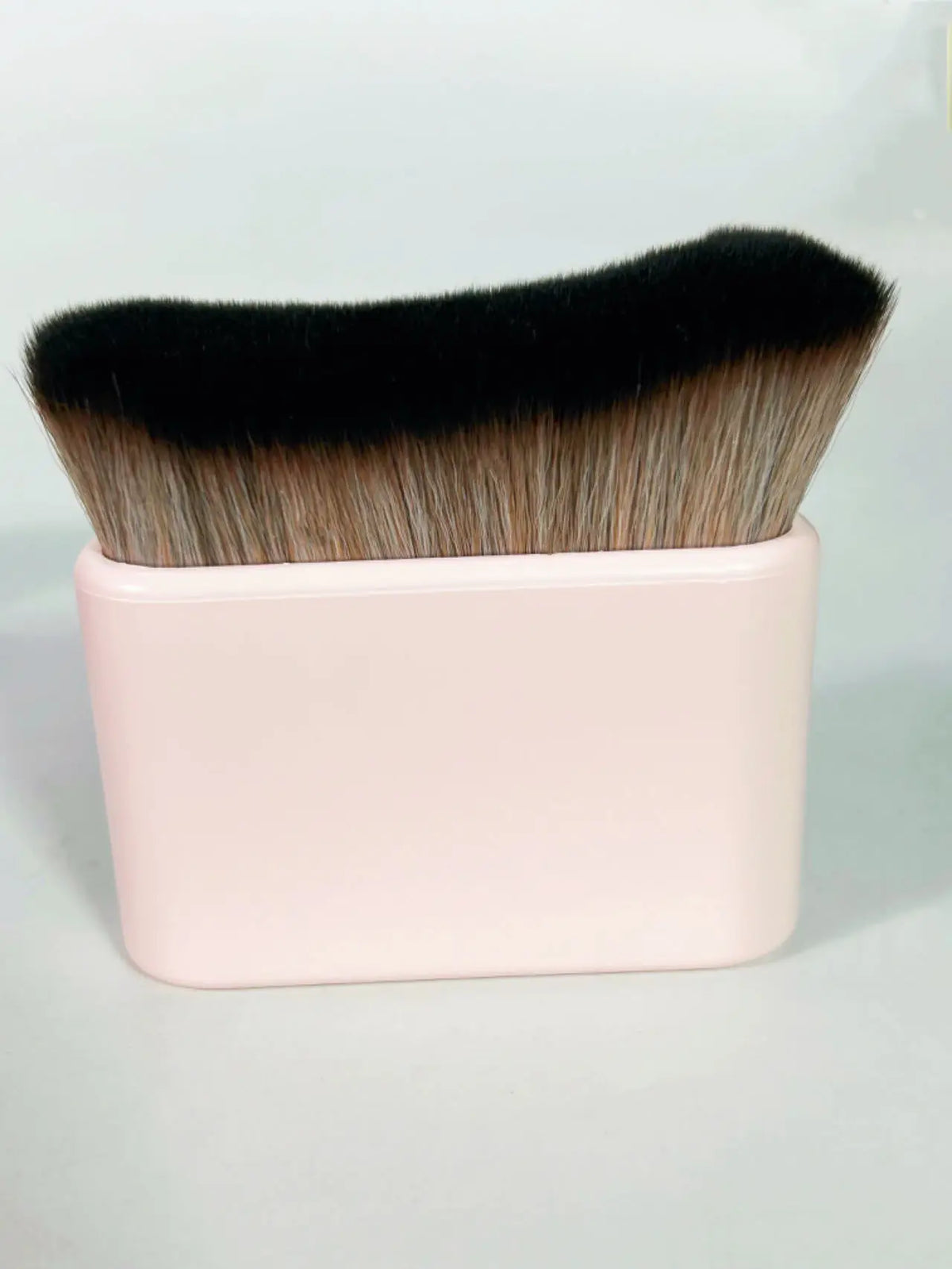 A makeup brush with black and tan bristles is shown in a pink rectangular container.