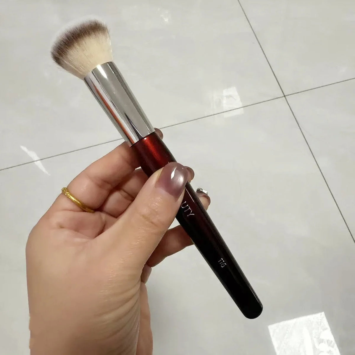 Une main tient un pinceau de maquillage liquide angulé avec une ferule argentée et une tête fluffy.