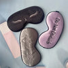 Three embroidered silk sleep masks in black, pink, and silver resting on a light blue surface.