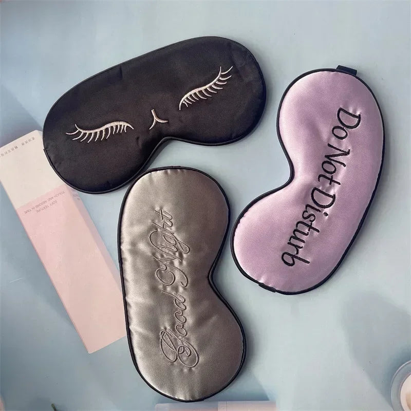 Three embroidered silk sleep masks in black, pink, and silver resting on a light blue surface.