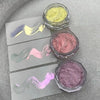 A collection of glitter powders in various shades including yellow, pink, and purple displayed in small glass jars.