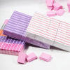A collection of pink and purple multi-sided nail and cuticle buffing files stacked and scattered on a white surface.