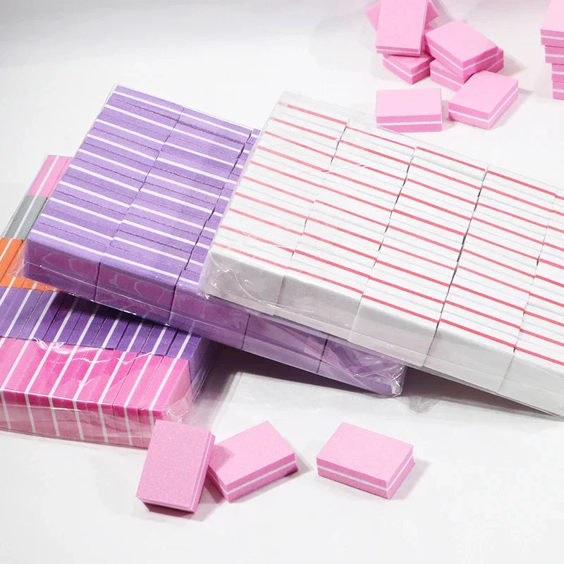 A collection of pink and purple multi-sided nail and cuticle buffing files stacked and scattered on a white surface.