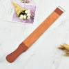 A brown leather sharpening strop with a metal clip rests on a white marble surface next to a small flower-themed card.