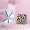 A silver double-sided portable makeup mirror is shown open and a leopard print compact case next to it on a pink surface.