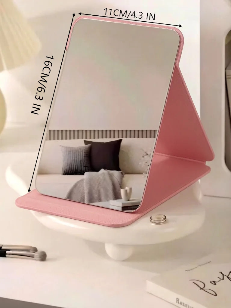 A pink foldable portable makeup mirror with measurements marked, resting on a white surface.