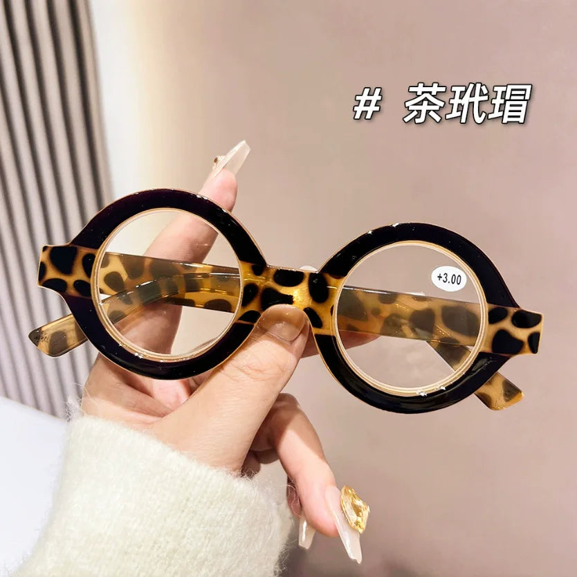 A hand holds a pair of round tortoiseshell acetate reading glasses with clear lenses against a light background.
