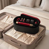 A black storage box containing a red hair styling tool and several small accessories, placed on a white bed.