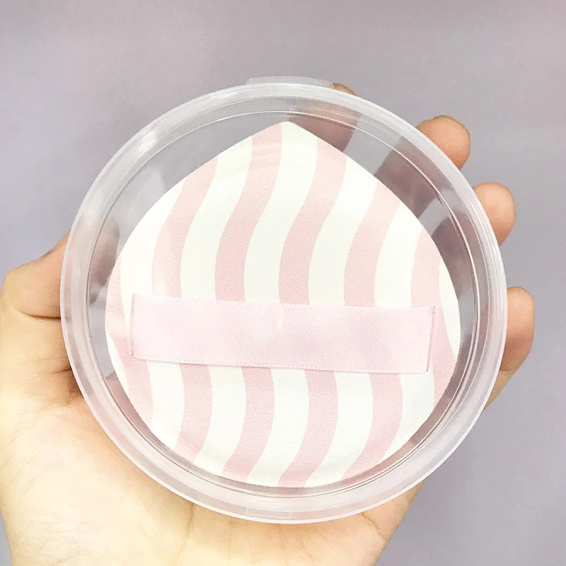 A pink and white striped makeup puff is visible inside a clear storage container being held in a hand.