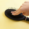 Eine Hand benutzt eine schwarze Kopfmassagebürste mit vielen Borsten auf einer hellgelben Oberfläche.