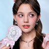 A young woman holds a pearl hand mirror for makeup and beauty, featuring a pink bow.