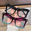A hand holds a pair of square-framed reading glasses with purple and blue tinted lenses.