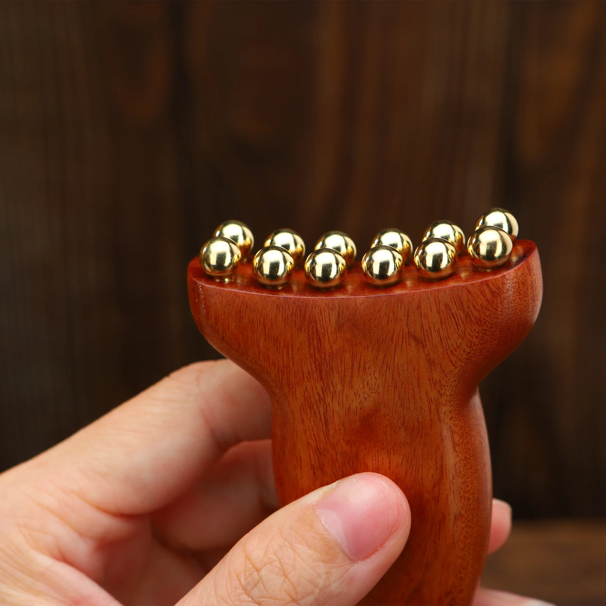 A hand holds a natural wood facial and scalp massage comb with gold-colored massage balls on the tips.