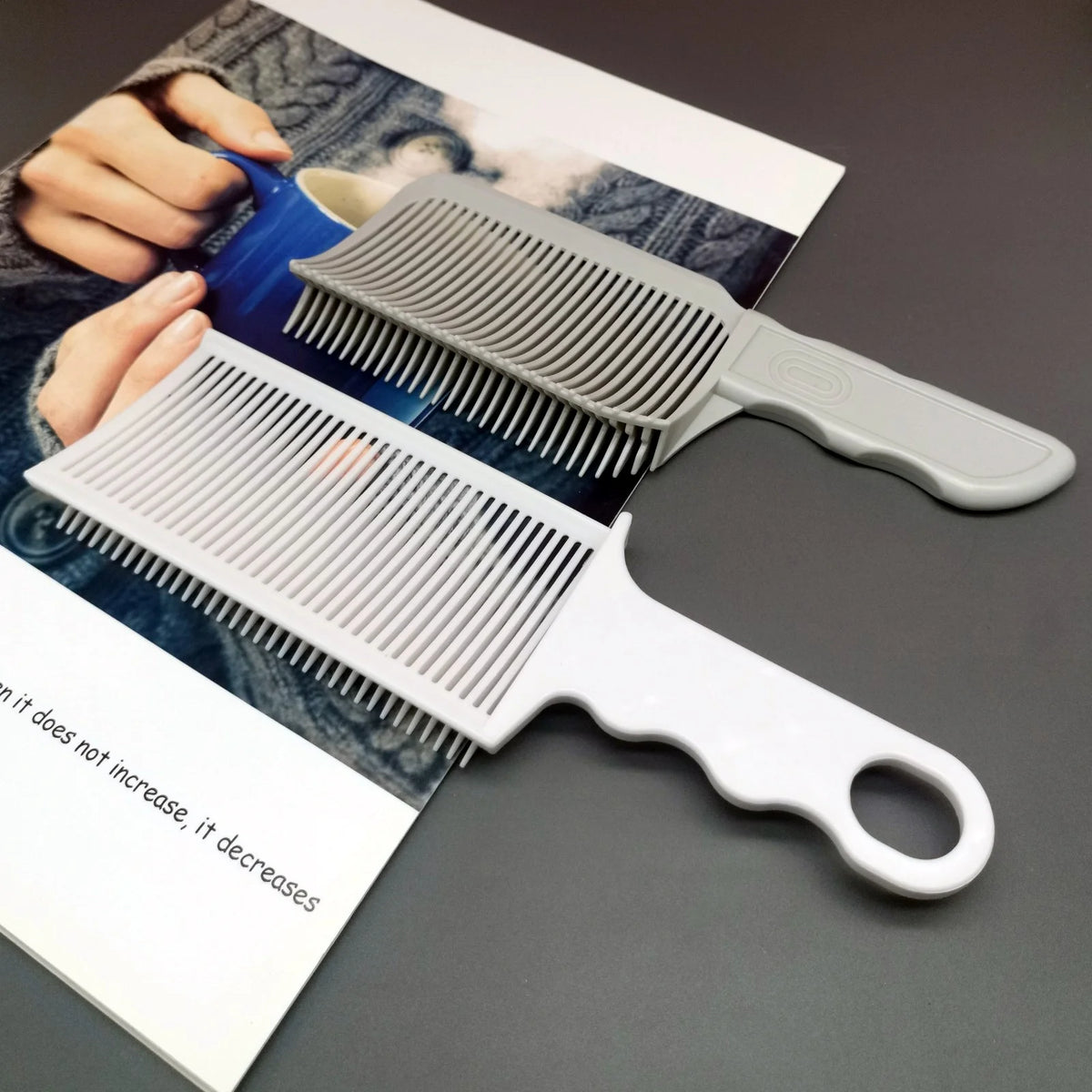 A silver-gray professional barber hair cutting comb is shown next to a white comb, with a hand holding a blue object in the b