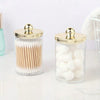 Distributeur en verre transparent avec couvercle doré contenant des cotons-tiges et des pompons blancs.