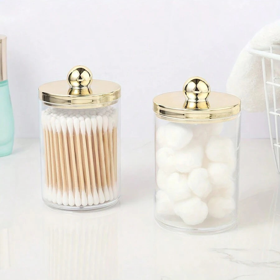 Distributeur en verre transparent avec couvercle doré contenant des cotons-tiges et des pompons blancs.