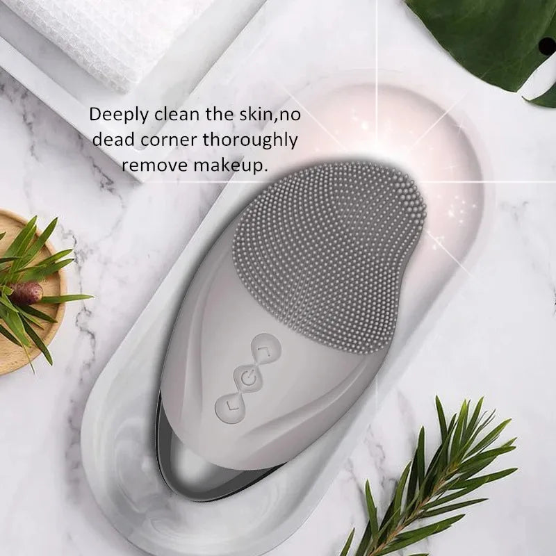 A USB rechargeable facial cleansing brush rests on a white tray next to green leaves.