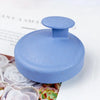 A smooth, light blue silicone scalp massage brush is shown resting on a white surface next to dried rose petals.