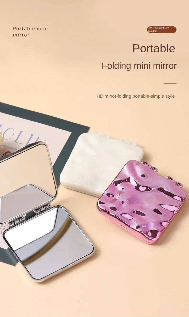 Un mini miroir pliable rose et violet à côté d'un chiffon de nettoyage blanc sur fond clair.