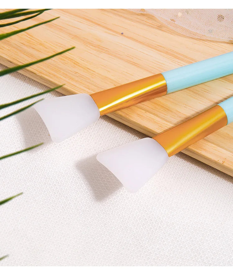 A silicone facial mask applicator brush with a gold and light blue handle rests on a wooden board.