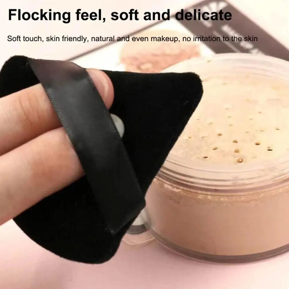 A hand holds a black triangular velvet powder puff next to a jar of light pink cosmetic cream.