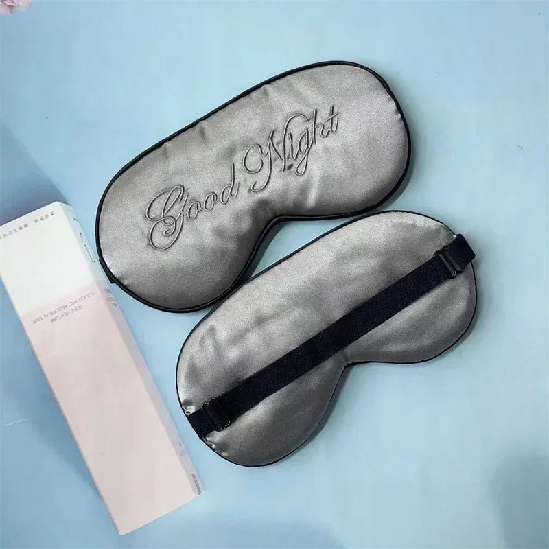 A gray silk sleep mask embroidered with the words Good Night is displayed next to its packaging.