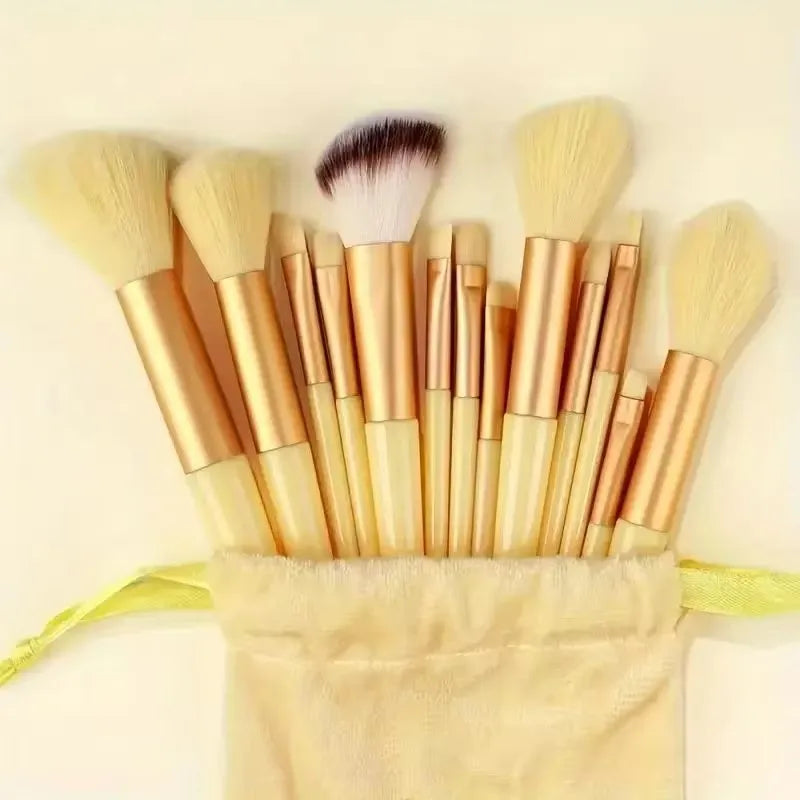 A collection of thirteen professional makeup brushes with golden handles and natural hair bristles, resting in a light yellow