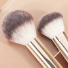 A pair of professional makeup brushes with silver and white bristles and light wood handles.