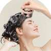 A woman is using a rechargeable electric handheld scalp massager on her hair, applying foam or lotion.