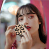A woman holds a black leopard print double-sided portable makeup mirror up to her face.