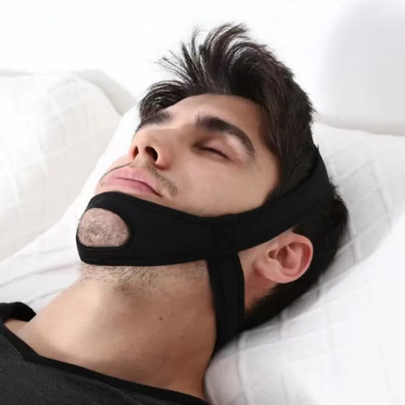 A man wearing a black anti-snoring chin strap while sleeping on a white pillow.