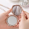 A hand holds an open vintage rose design double-sided pocket makeup mirror with a silver-toned frame.
