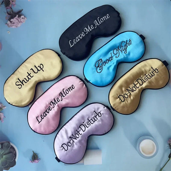 A collection of embroidered silk sleep masks in black, light blue, pink, and gold tones arranged on a light blue surface.