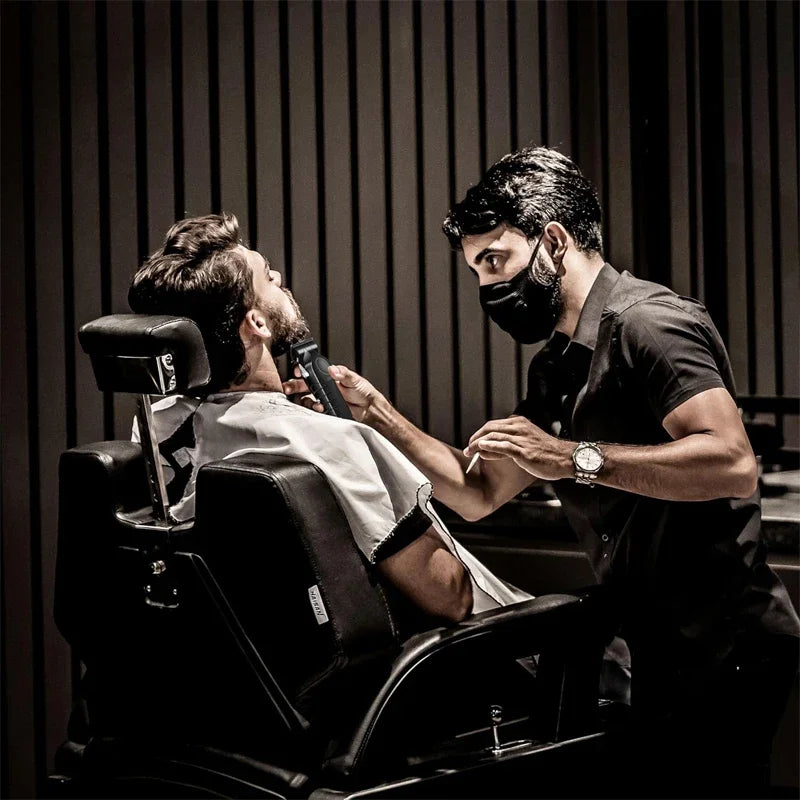 A man is using professional cordless electric hair clippers on another man's beard.