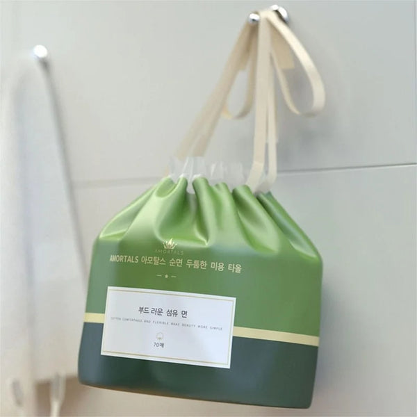 A green pouch of soft cotton disposable face towels with a light beige tie, hanging on a white surface.