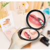 A hand holds a portable magnifying makeup mirror showing a close-up of pink lipstick on lips.