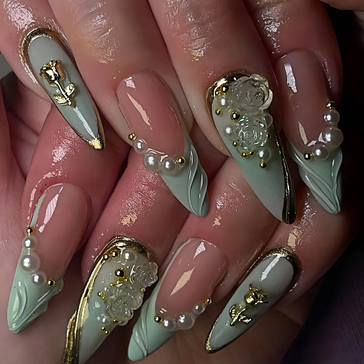A close-up of a hand displaying 24-piece almond press-on nails featuring floral and butterfly designs in pale green and gold.