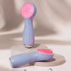 A light blue vibrating LED facial cleansing brush resting on a white circular stand.