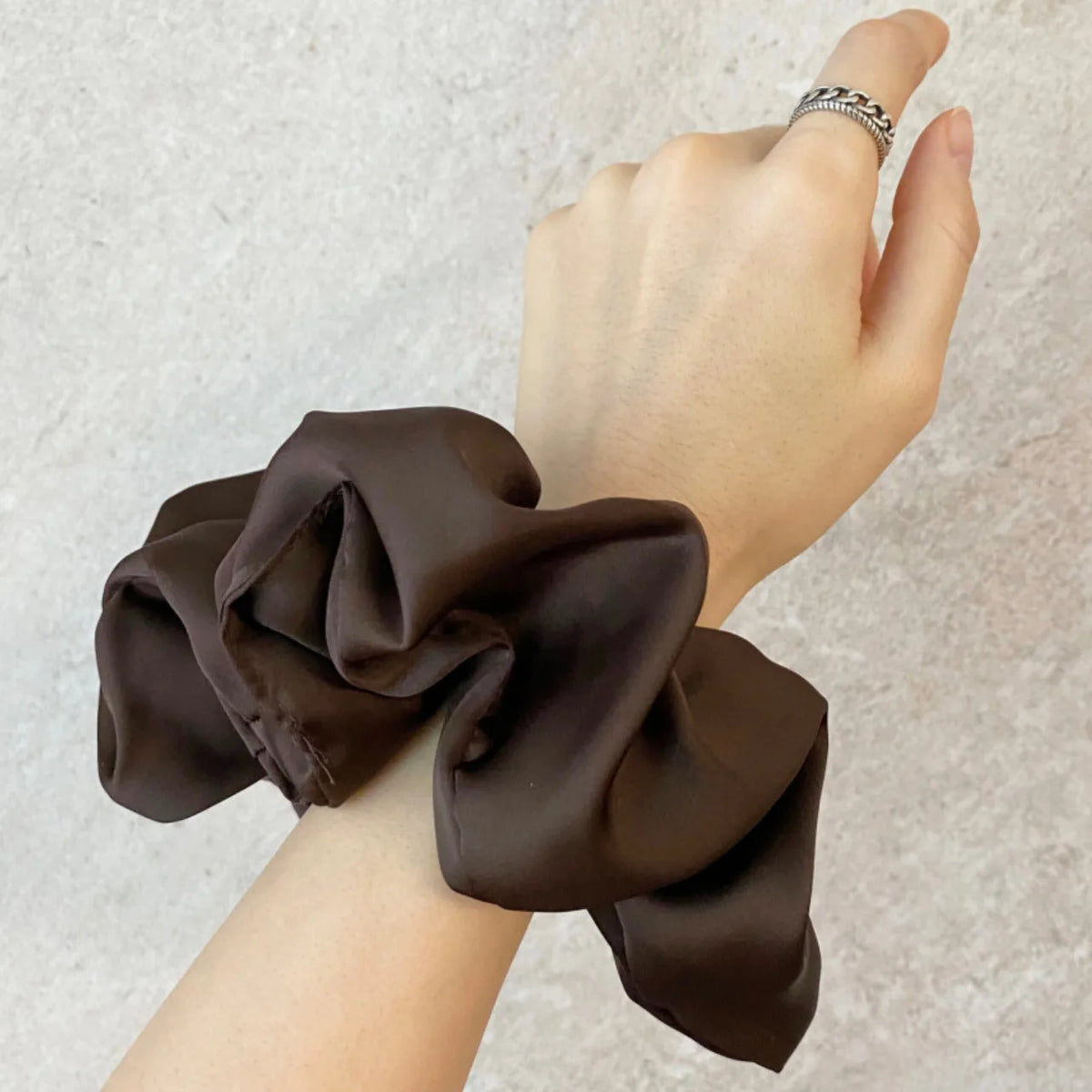 Une main porte un élastique pour cheveux en mousseline de satin brun foncé au poignet.