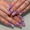 A hand displays purple and white press-on nails with a floral wave design on a light background.