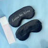 Black silk sleep mask embroidered with the text Leave Me Alone resting on a light blue surface.