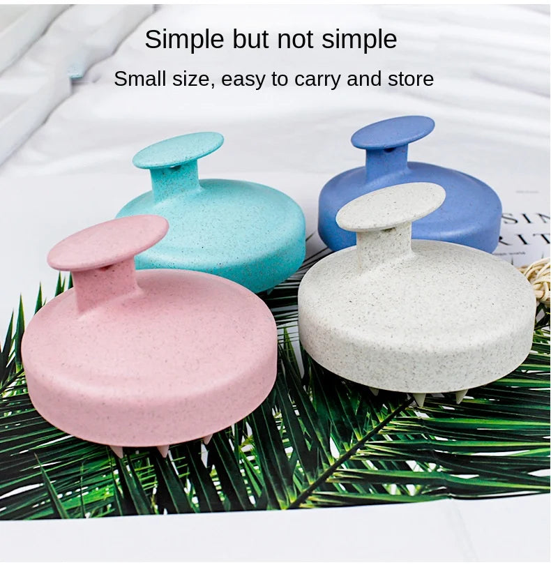 Three colored silicone scalp massage brushes in pink, light blue, and gray resting on green leaves.