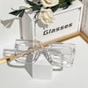 A pair of clear square eyeglasses resting on a white block next to a box with the word Glasses.