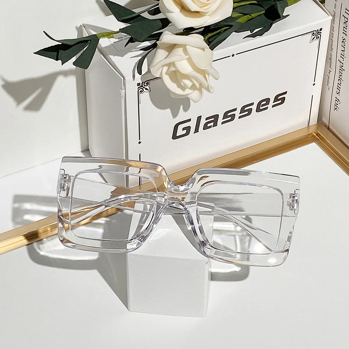 A pair of clear square eyeglasses resting on a white block next to a box with the word Glasses.