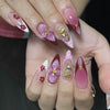 Eine Hand zeigt ein Set vergoldeter Mandelblüten-Press-On-Falschnägel mit rosa und lila Akzenten.