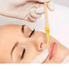 Une main utilise un bâton de dépilation en bois naturel pour enlever les poils de l'arcade sourcilière d'une femme.