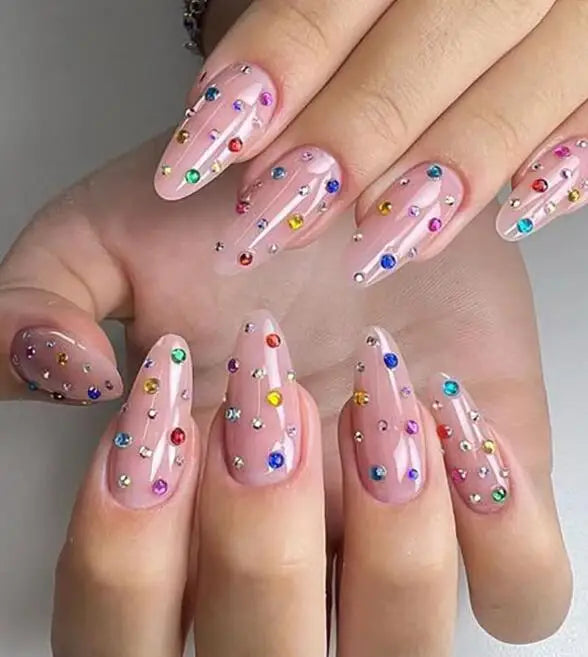 Une main montrant de longues ongles roses et irisants décorés de strass colorés et de paillettes.