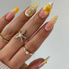 A hand displaying various almond press-on nails featuring floral and butterfly designs in yellow, white, and gold accents.
